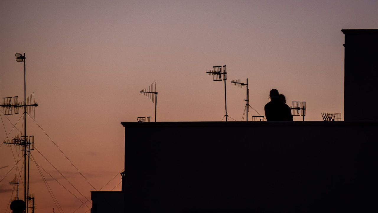 Rooftop TV antennas and masts silhouetted against an orange and purple sunset, with two small figures on a roof terrace